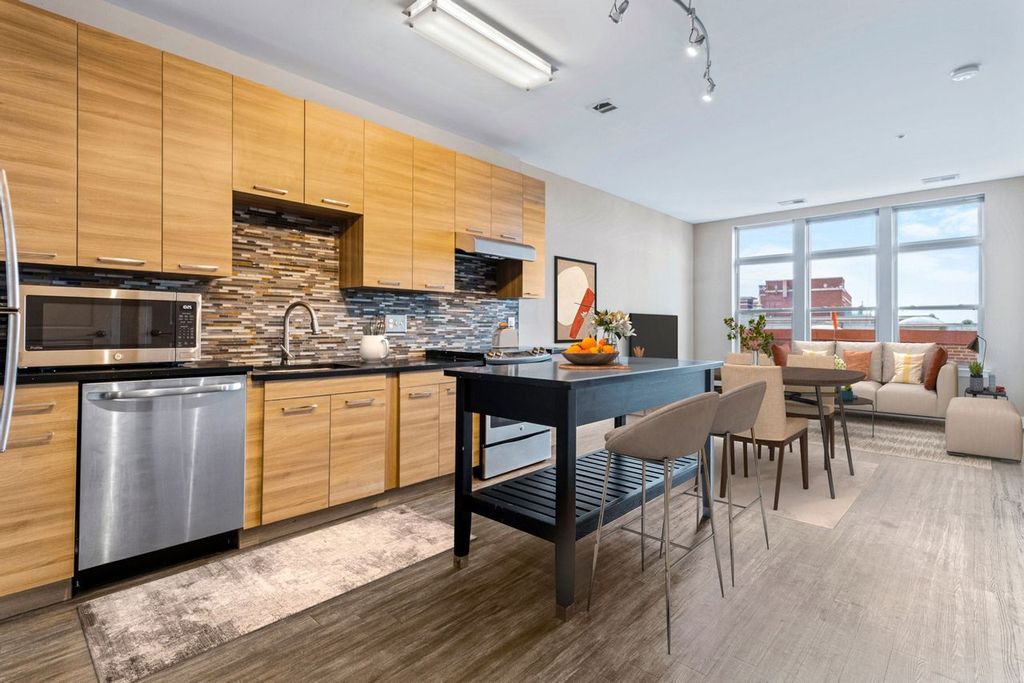 Modern open-concept kitchen and living area at The Swift at Petworth Metro, featuring wood cabinets, stainless steel appliances, island, dining table, and large windows letting in natural light. Eligible for the IZ Housing Program.