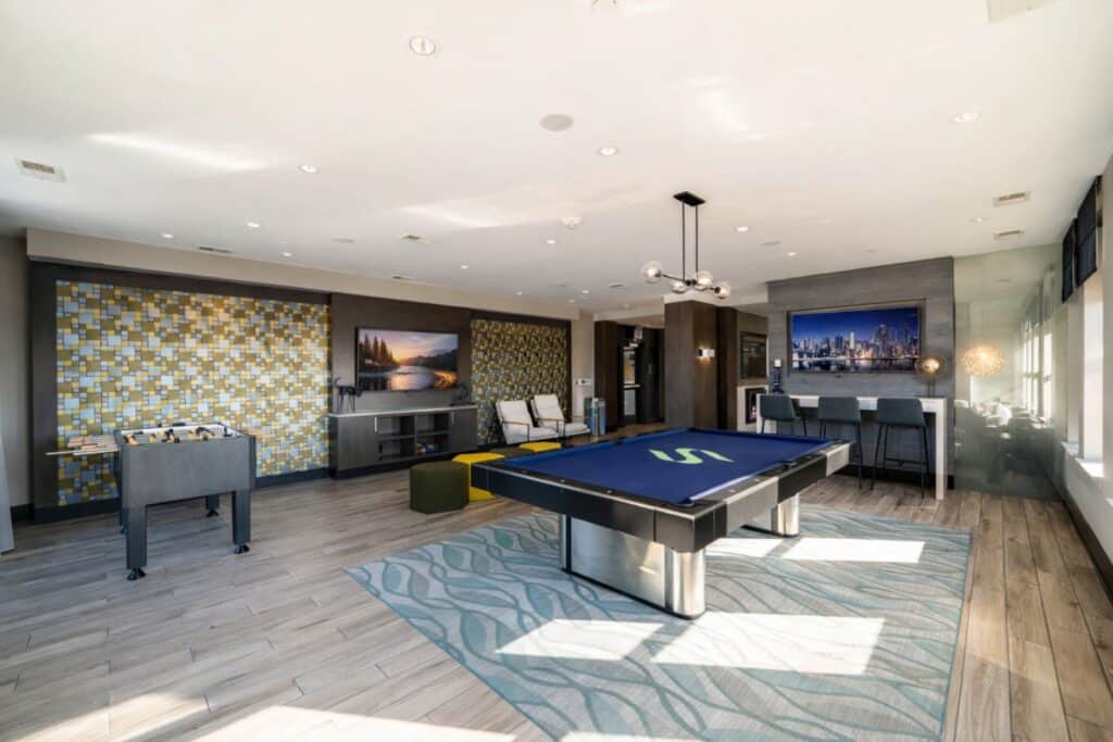 Modern recreation room at Swift at Petworth features a foosball table, blue pool table, lounge seating, bar area, large windows, and wall-mounted TVs.